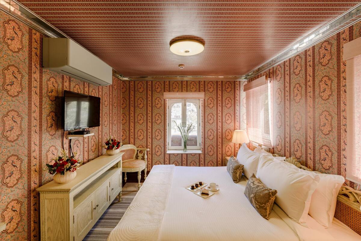 A hotel room with a large bed, a TV on the wall, a window with curtains, a dresser with a vase of flowers, and a small table with a tray of snacks.