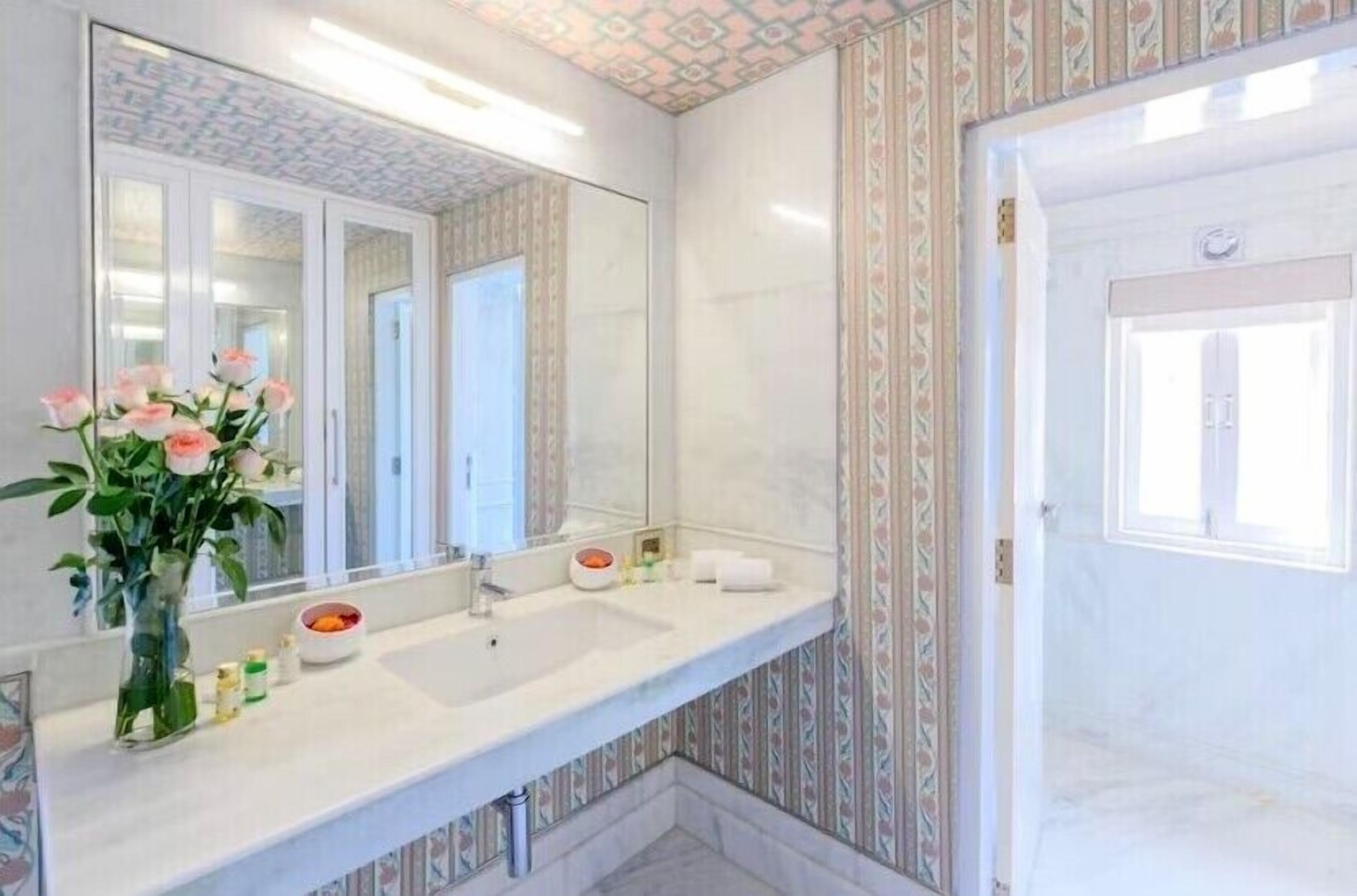 A bathroom with a large mirror, two sinks, a vase with flowers, and patterned wallpaper.
