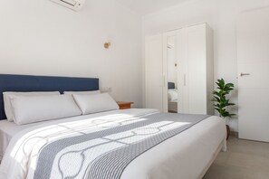 A neatly made bed with white linens and a blue headboard, a bedside table with a lamp, and a potted plant in a corner.
