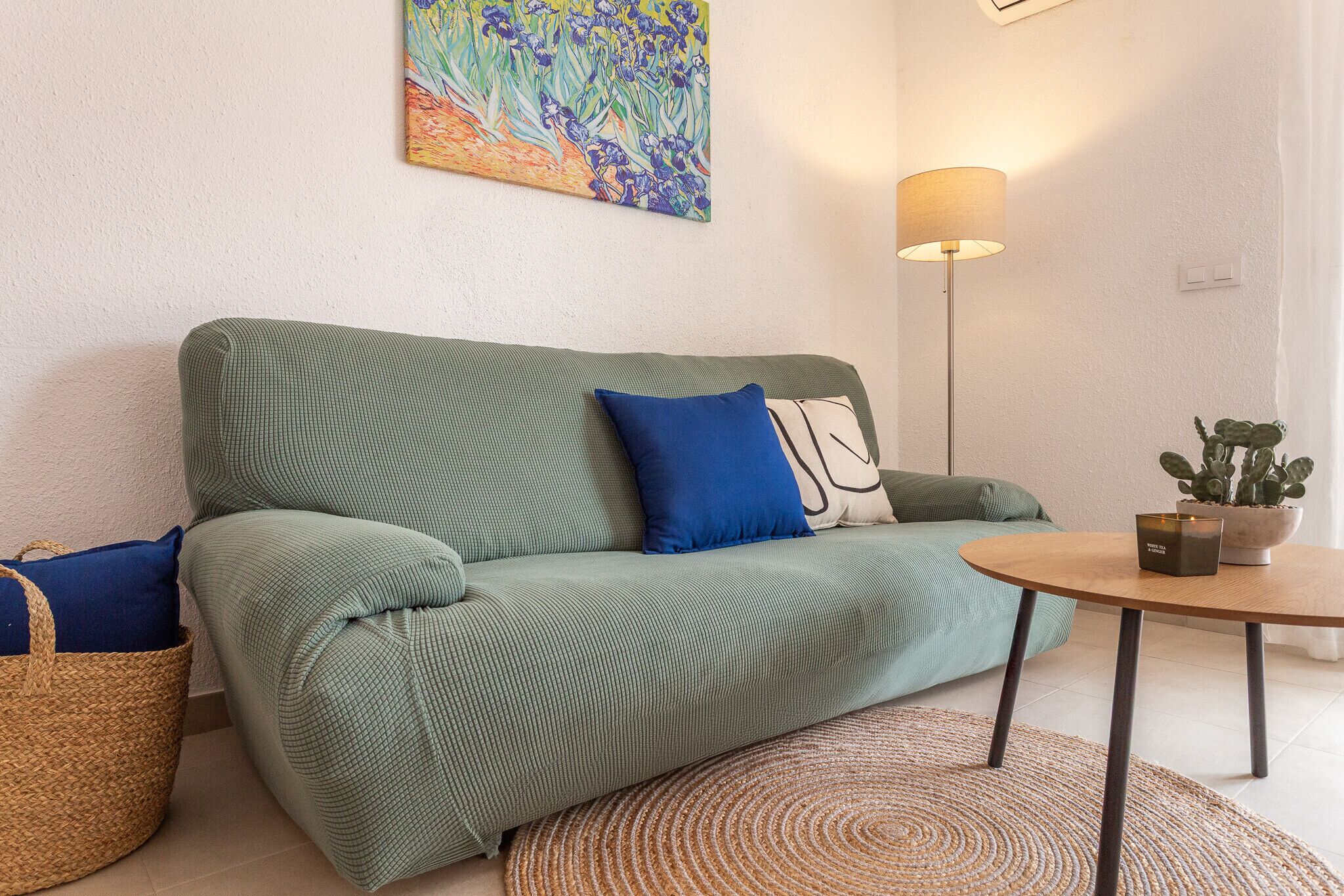 A green sofa with blue and white cushions, a round wooden table with a potted plant, and a floor lamp.