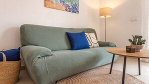 A green sofa with blue and white cushions, a round wooden table with a potted plant, and a floor lamp.