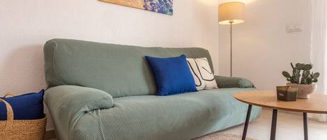 A green sofa with blue and white cushions, a round wooden table with a potted plant, and a floor lamp.