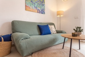 Een groene bank met blauwe en witte kussens, een ronde houten tafel met een plant in pot, en een vloerlamp.