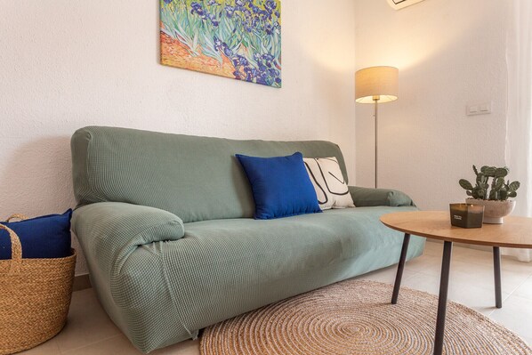 A green sofa with blue and white cushions, a round wooden table with a potted plant, and a floor lamp.