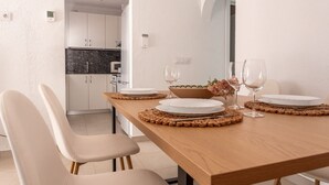 A dining area with a wooden table set for two, white chairs, and a kitchen in the background.