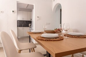 A dining area with a wooden table set for two, white chairs, and a kitchen in the background. - Casa Iris only 700 meters away from the beach (L'Escala)