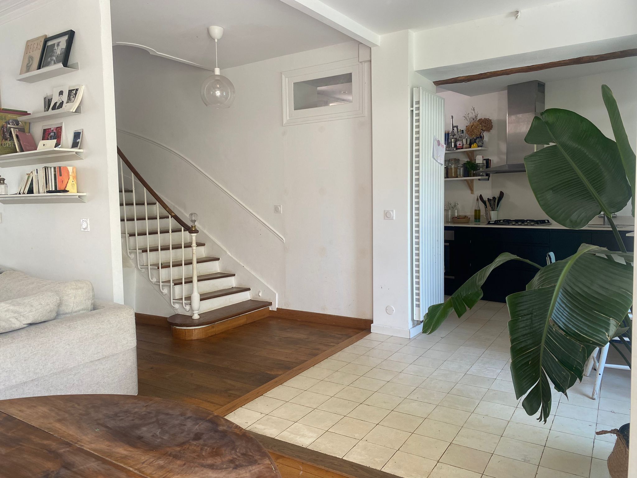 Een ruime woonkamer met een trap, een keuken die zichtbaar is door een open deur, en een grote groene plant.