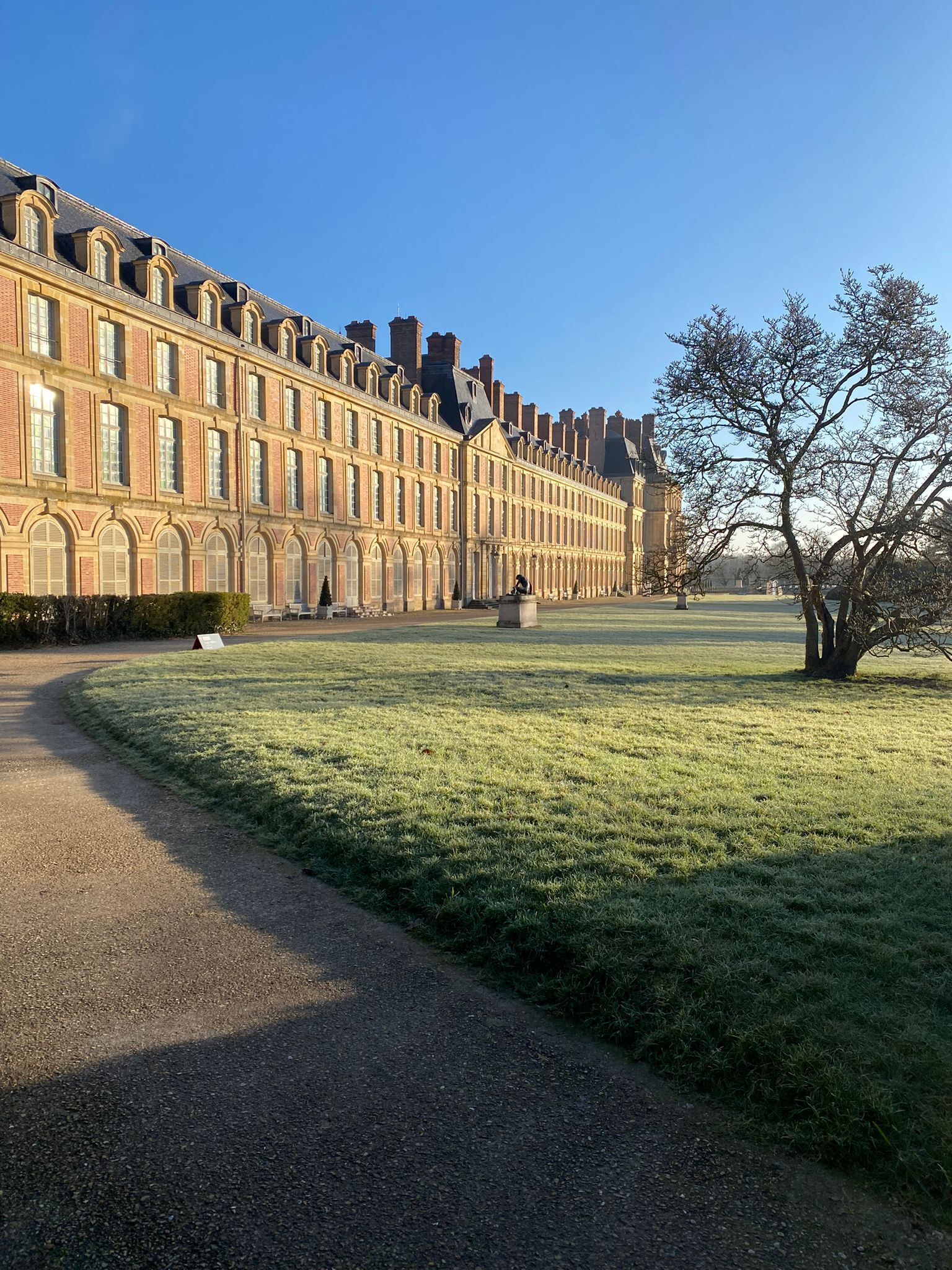 A large, historic building with a symmetrical facade, a central courtyard, and a gravel path leading to the entrance.