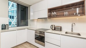 Une cuisine moderne avec des armoires blanches, un four encastré, un évier en acier inoxydable et une étagère en bois au-dessus du plan de travail.