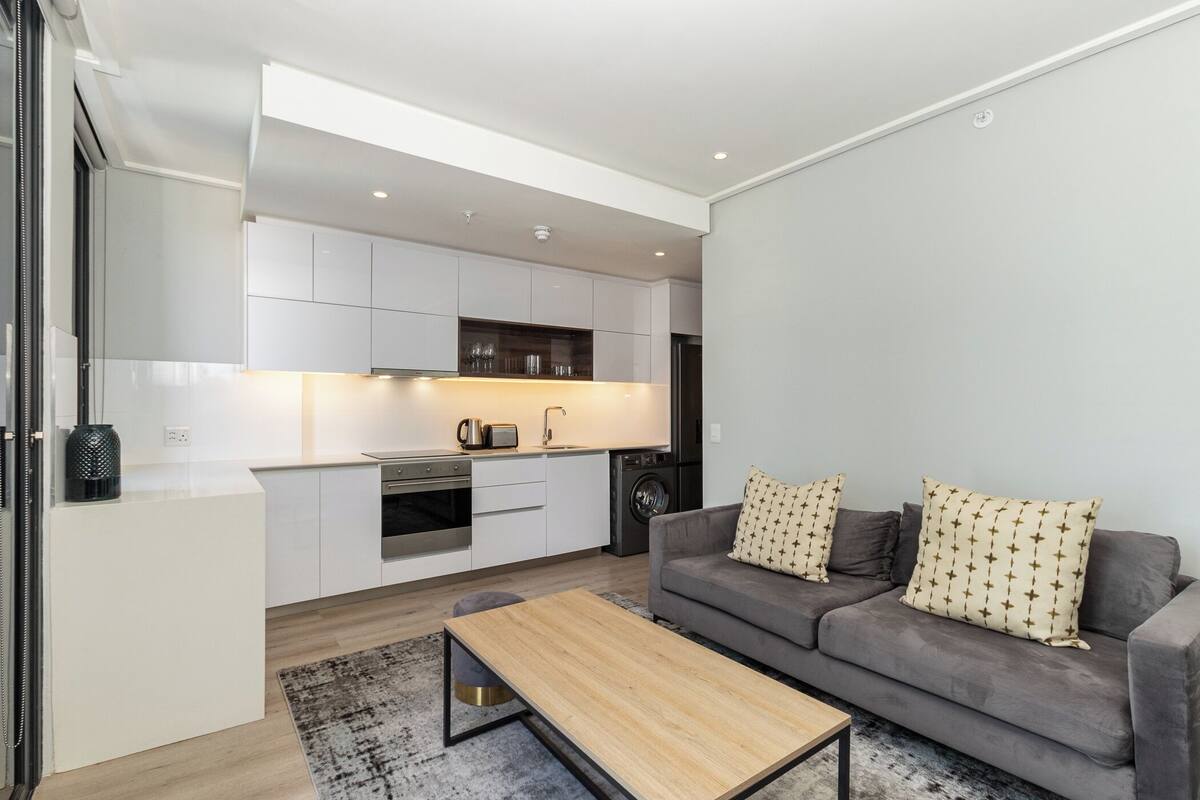 A modern apartment with a kitchen, a grey sofa, and a wooden coffee table.