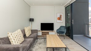 Un salon moderne avec un canapé gris, une table basse en bois, un téléviseur à écran plat posé sur un meuble en bois, un fauteuil bleu et une grande fenêtre donnant sur des immeubles.