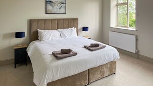 A neatly made bed with white linens and two folded towels, a large abstract painting on the wall, and a window with a view of greenery.