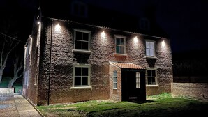 A brick house with multiple windows and a door, illuminated by exterior lights at night.