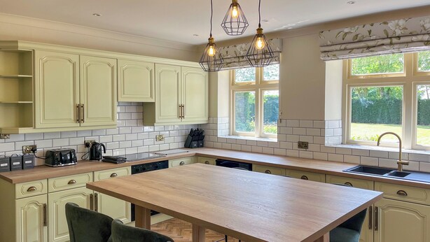 A kitchen with light green cabinets, a wooden island with bar stools, a window with a view of greenery, and pendant lights.