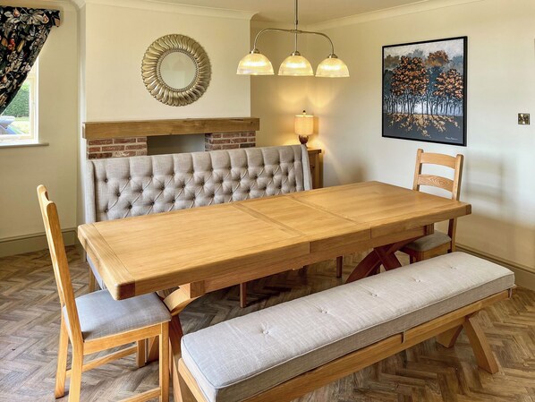 A dining area with a wooden table, cushioned bench, and chairs. A brick fireplace is visible in the background.