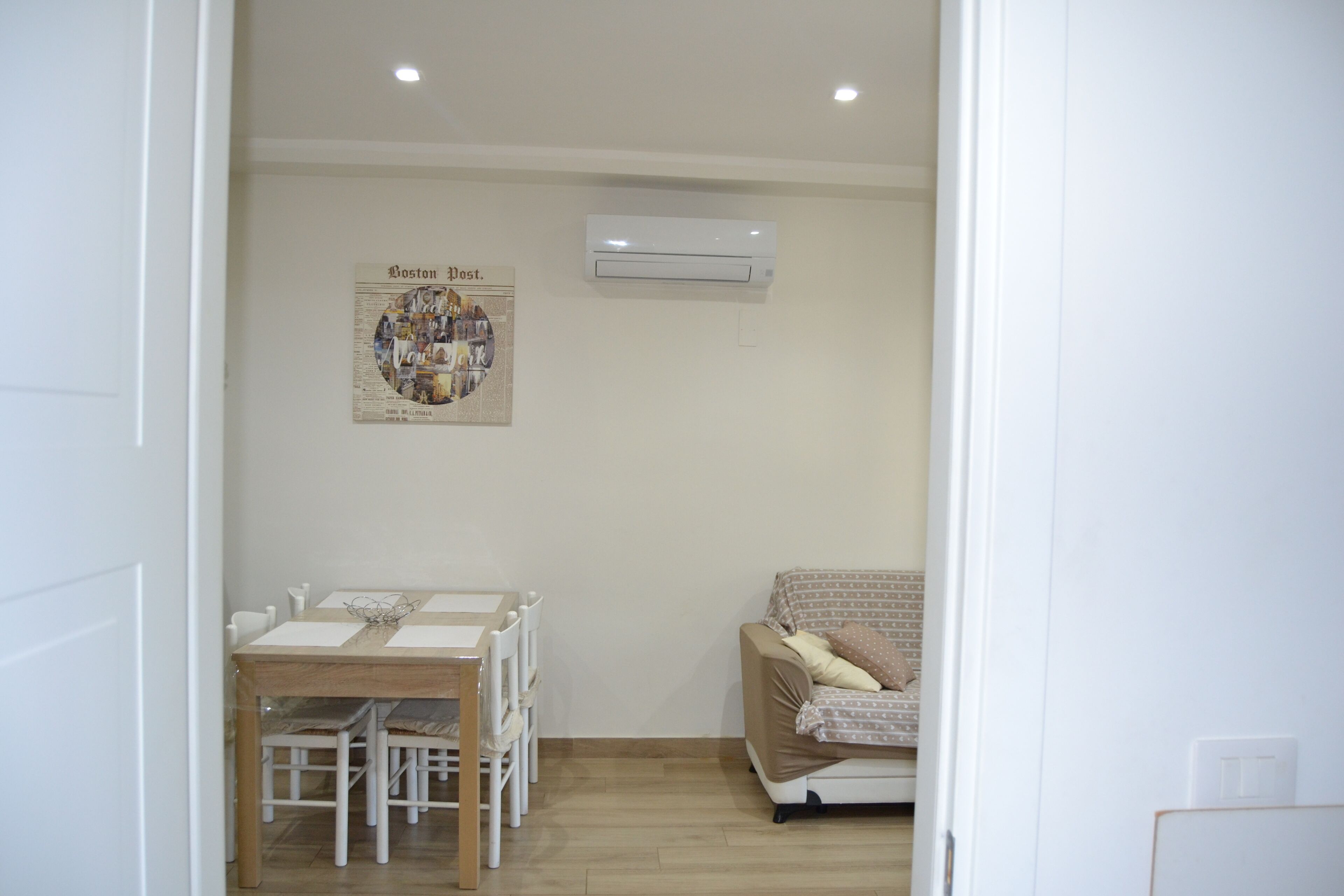 A room with a dining table and chairs, a sofa, an air conditioning unit, and a wall-mounted poster.