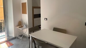Una habitación pequeña y moderna con una mesa blanca y sillas, una mesita auxiliar con una botella y un vaso, y vistas a un balcón a través de una puerta corredera.