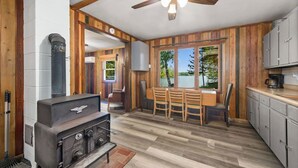 A rustic cabin interior with a wood-paneled kitchen, a dining area with a view of a lake, and a large stove. - Turtle Lake Beach House - Get Up North Retreats (Bigfork)