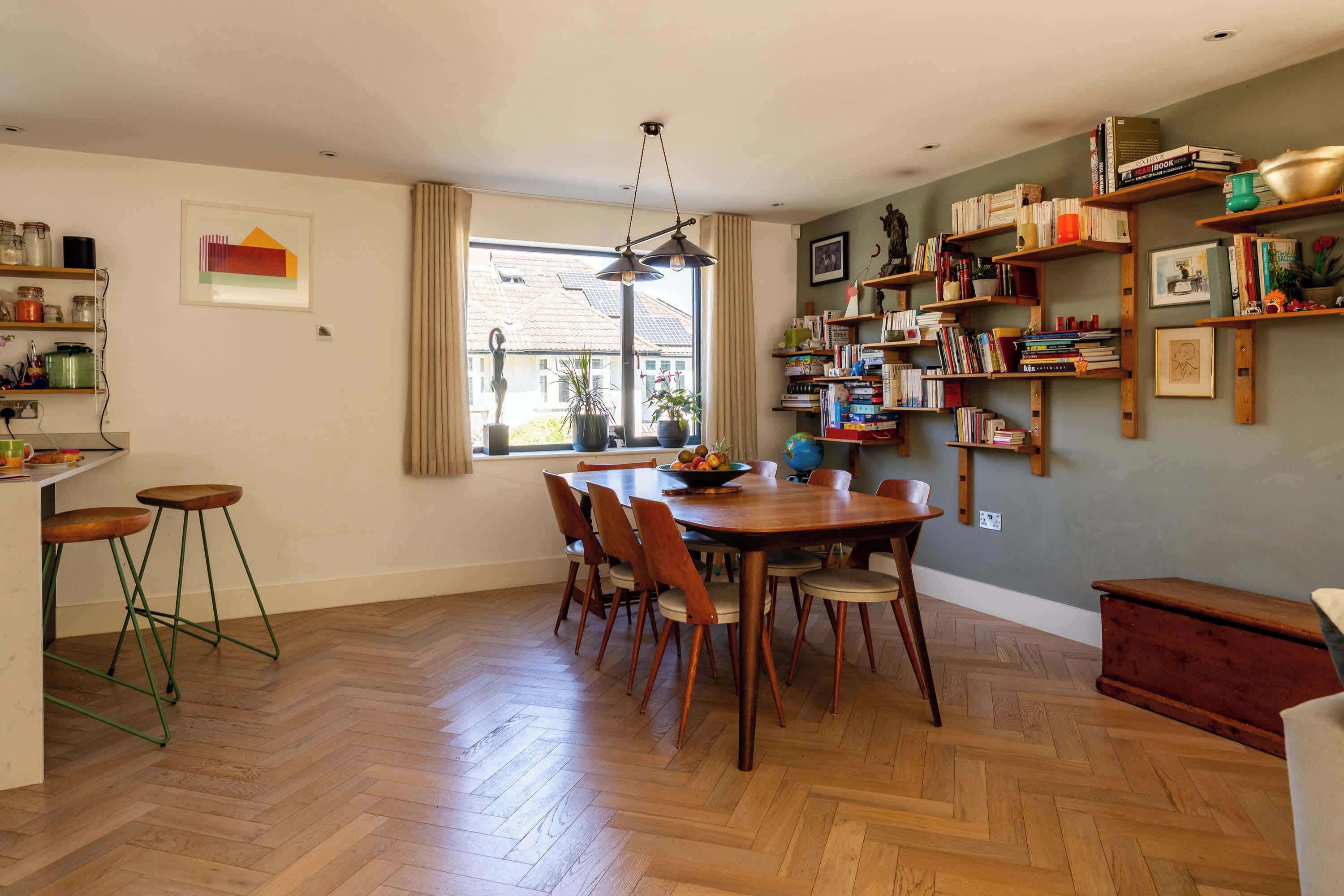 Uma área de refeições com mesa e cadeiras de madeira, cercada por estantes de livros e uma janela com cortinas.