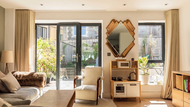 A modern living room with a sofa, armchair, coffee table, and a large mirror. There is a sliding glass door leading to a balcony with plants and a view of the outside. - Stylish Family Home with Garden (Bristol)