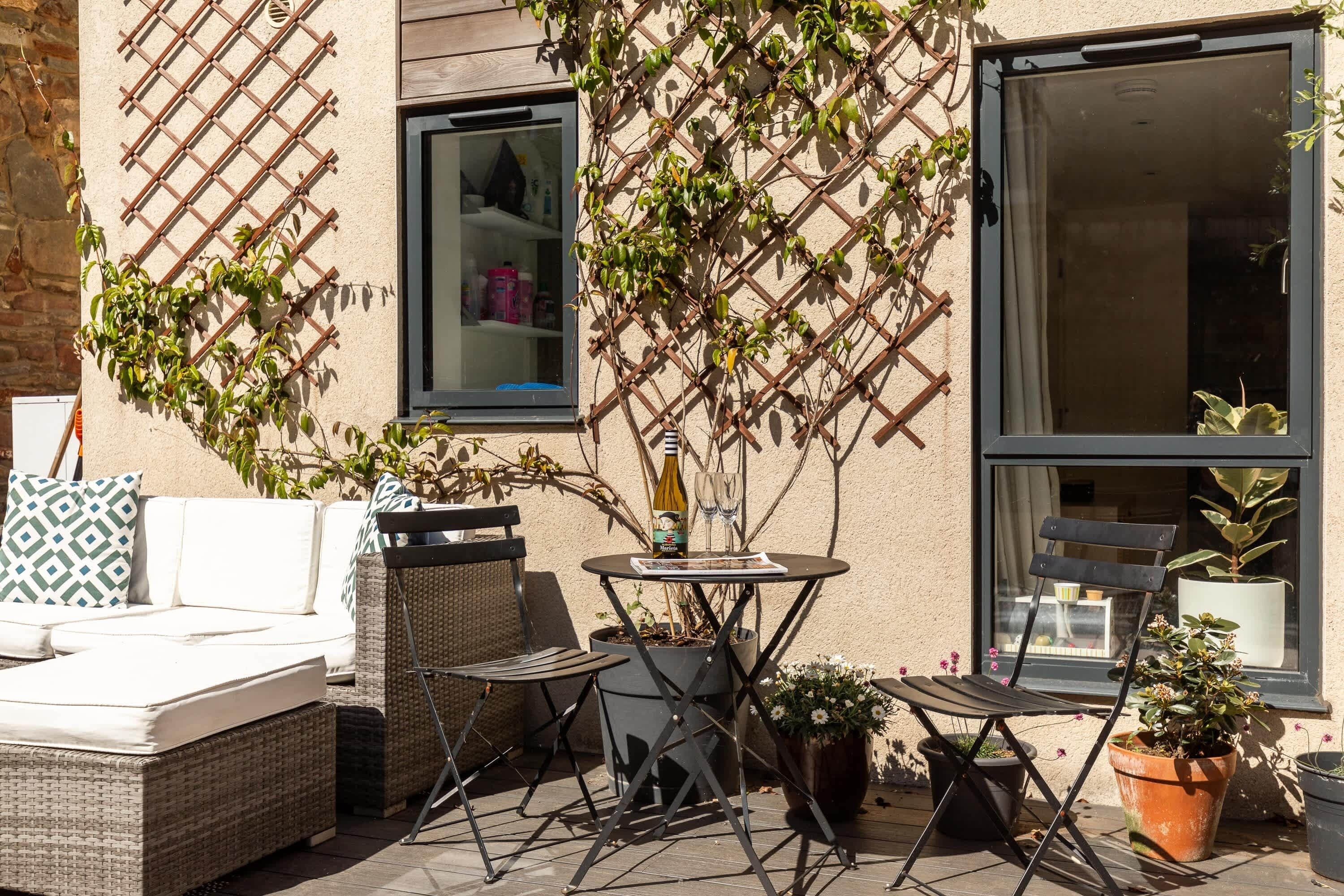 Een terras met een rieten bank, een klein rond tafeltje en een klapstoel. Er staan potplanten en een raam met uitzicht op het interieur.