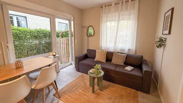 Una sala de estar con un sofá, una mesita con una planta, un comedor con sillas y vistas al exterior con vegetación.