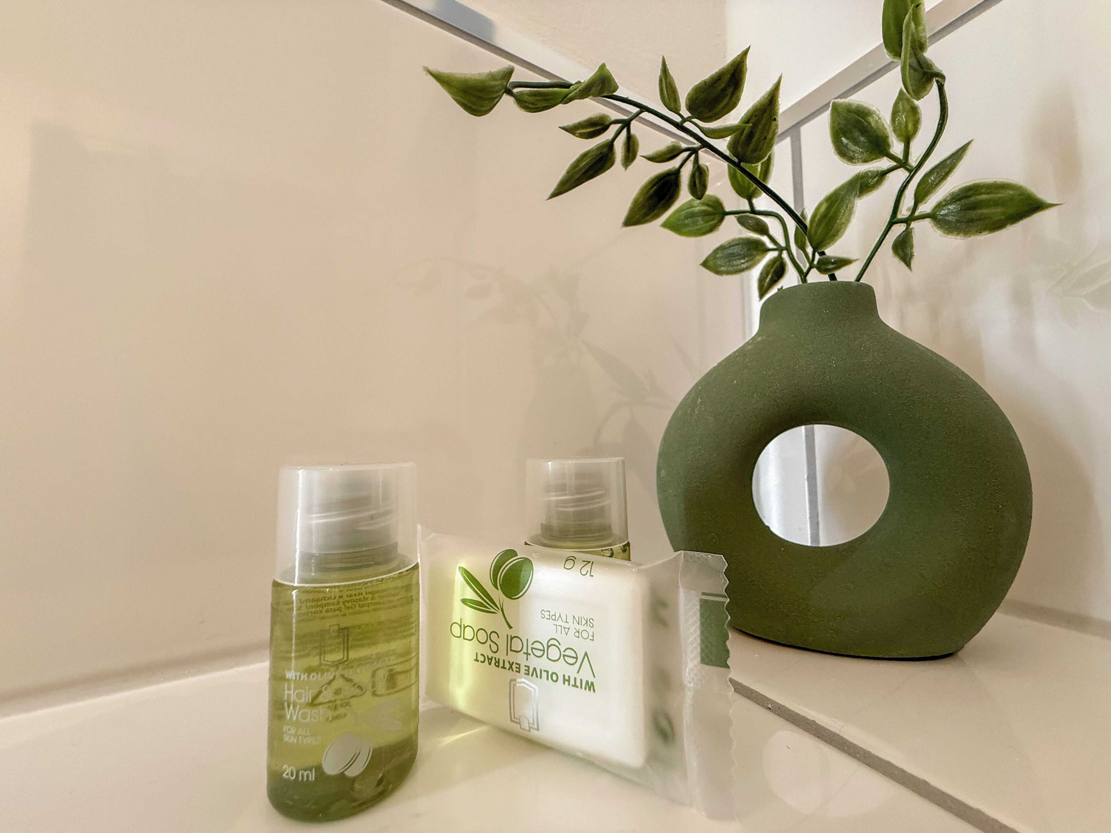 Une étagère de salle de bain avec un vase vert, deux flacons de produits de soins pour les cheveux et la peau, et un verre transparent.