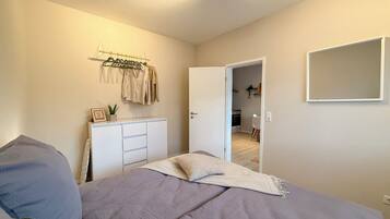 A bedroom with a bed, a bedside table, a hanging clothes rack, and a door leading to a kitchen.