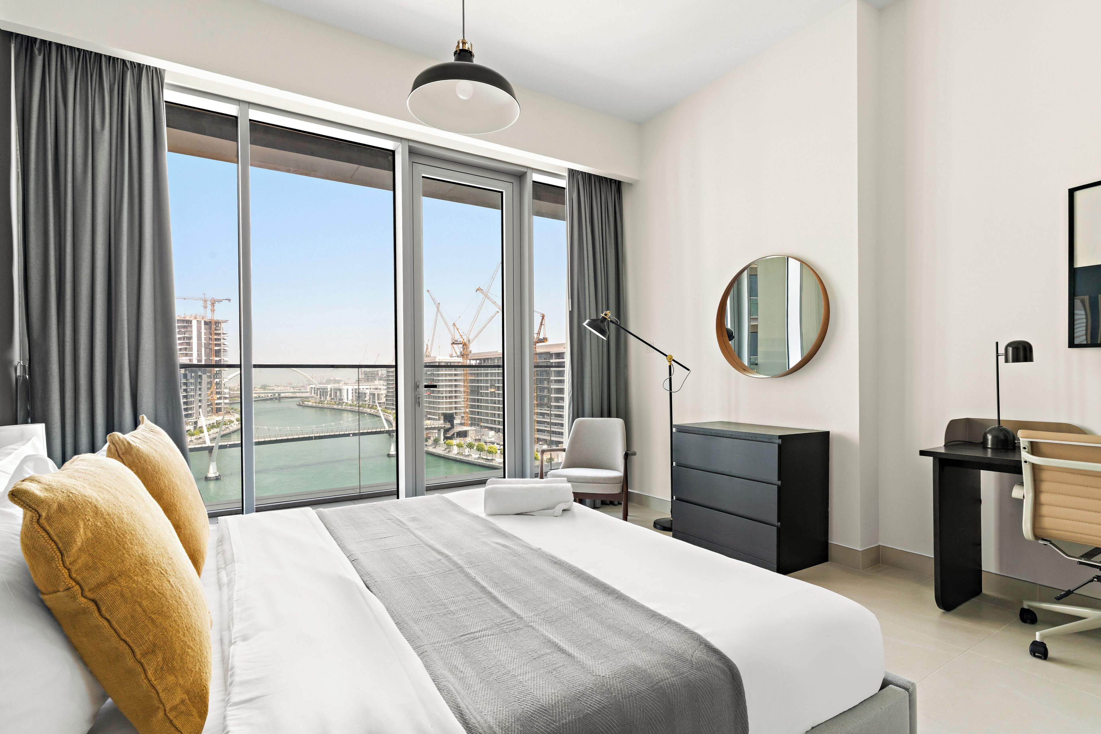 A modern hotel room with a large bed, a desk, a chair, a desk lamp, a round mirror, and a view of a cityscape through floor-to-ceiling windows.