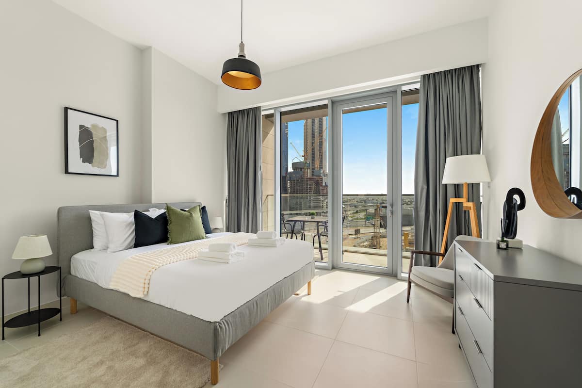 Un dormitorio moderno con una cama grande, un escritorio, una silla, una lámpara, un cuadro enmarcado y vistas de un paisaje urbano a través del balcón.
