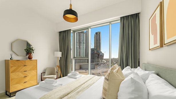 A hotel room with a large bed, a dresser, a chair, a desk, a lamp, and a view of tall buildings. - Silkhaus Canal Front 9 A tower (Dubai)
