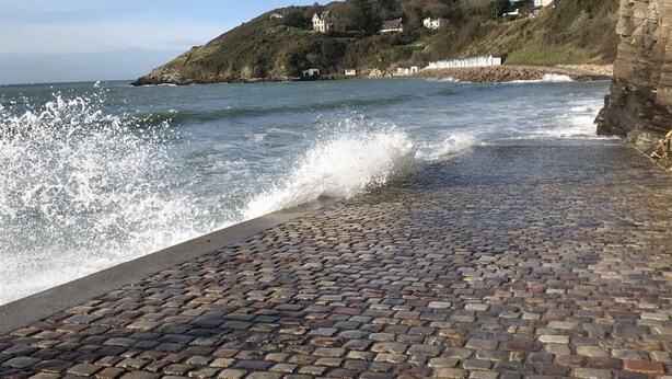 Un paseo empedrado junto al mar, con olas rompiendo en la orilla, un faro al fondo y una colina con edificios.