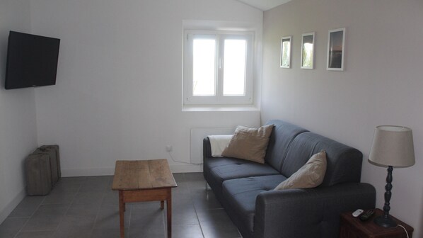 A living room with a grey sofa, a wooden table, a TV mounted on the wall, and a window with blinds.