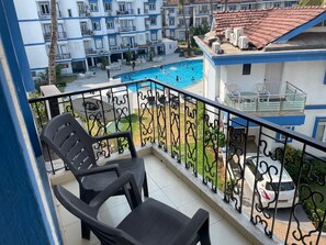 A balcony with two black plastic chairs, a pool, and a view of a residential area with buildings and a car. - Mariana By the Pool Benaulim (Benaulim)