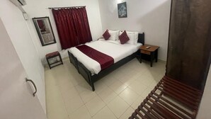 Un dormitorio con cama, mesita de noche, ventilador de techo, ventana con cortinas y un cuadro colgado en la pared.