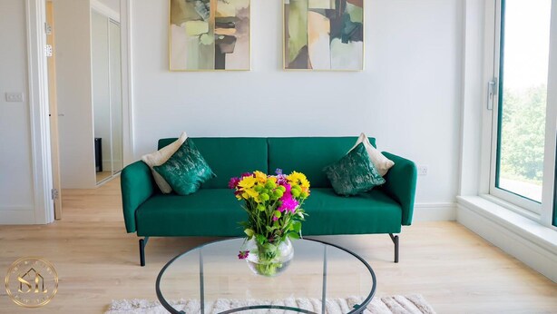 A living room with a green sofa, a glass coffee table with a vase of flowers, abstract paintings on the wall, and a view through a large window.