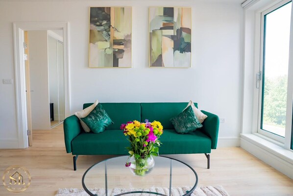 Ein Wohnzimmer mit einem grünen Sofa, einem Glastisch mit Blumen und abstrakten Bildern an der Wand, sowie einer Aussicht durch ein großes Fenster.