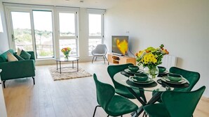 A modern living room with a dining area, large windows, a flat-screen TV, and a green sofa.