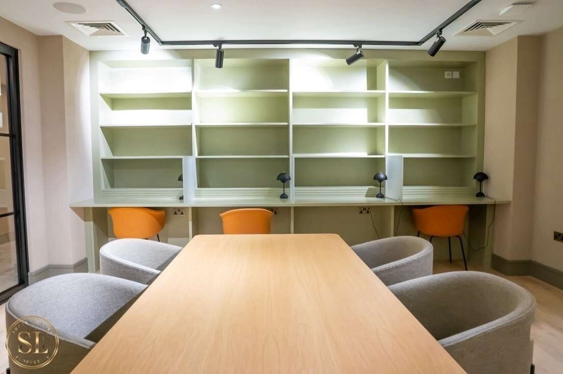 A conference room with a long table, four chairs, and a shelving unit with multiple compartments.