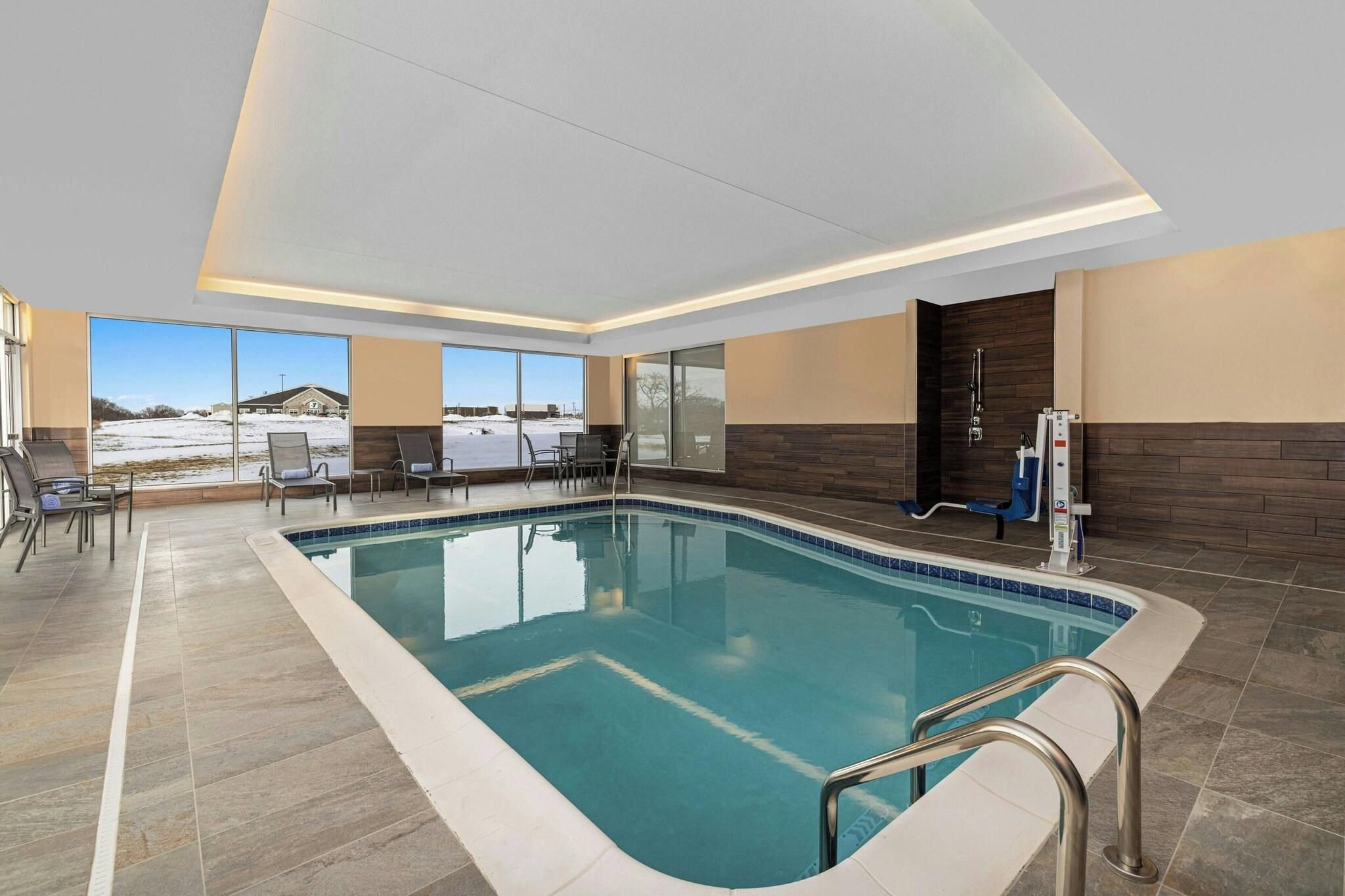 A modern indoor swimming pool with clear glass walls, a view of a snowy landscape, and a seating area with chairs.