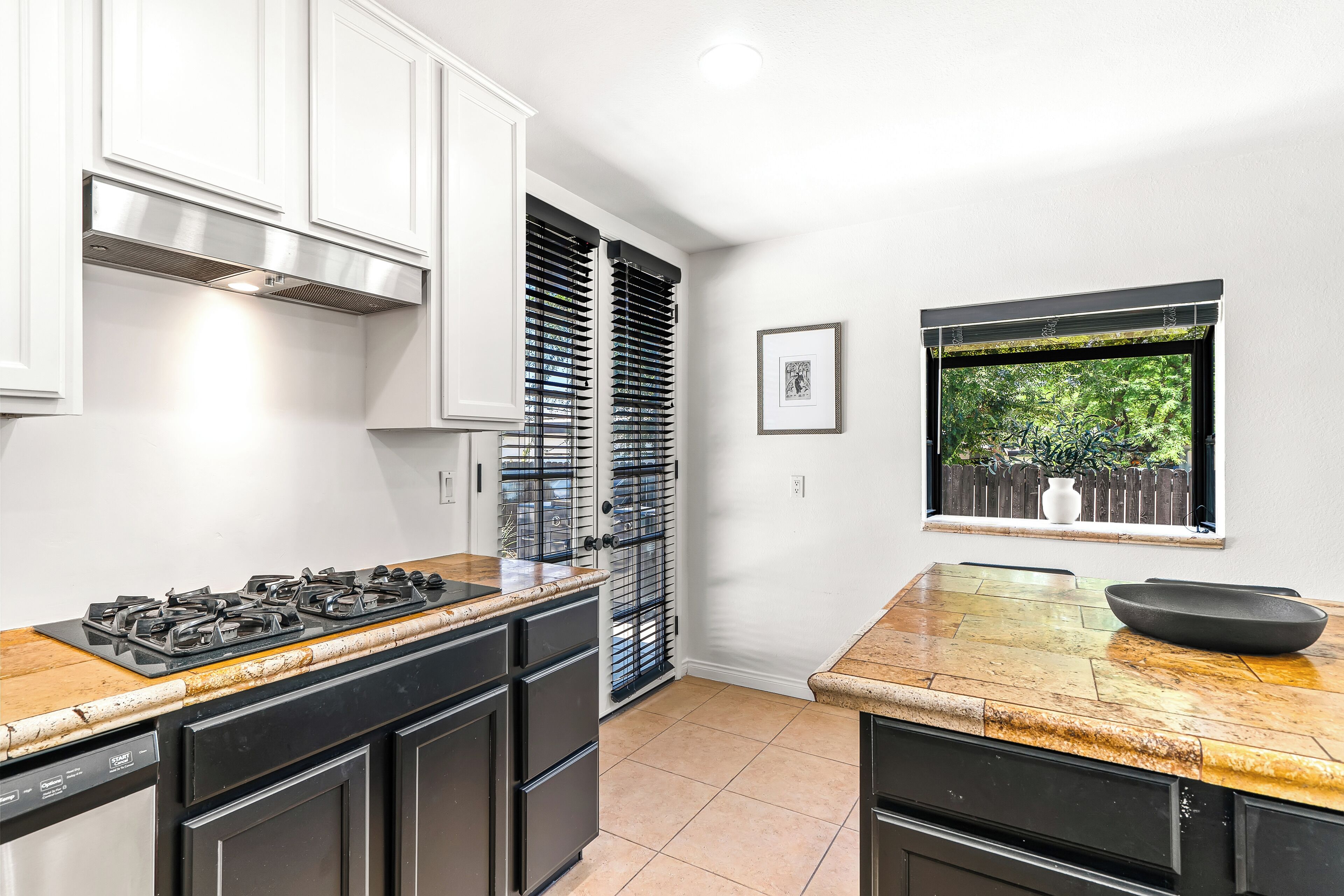 A kitchen with a gas stove, black cabinets, a window with a view of greenery, and a framed picture on the wall.
