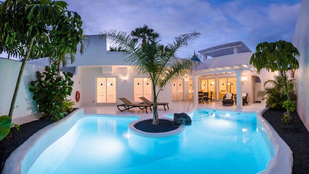 Un área de piscina moderna con sillones de descanso y una palmera, rodeada por un edificio blanco con puertas de vidrio y un porche cubierto.