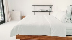 Un dormitorio minimalista con estructura de cama de madera, ropa de cama blanca, una mesita de noche pequeña y un estante con una cámara y objetos decorativos.