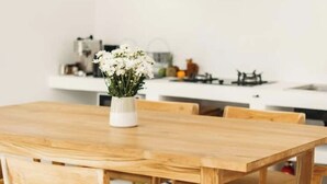 Una mesa de madera con sillas, un jarrón con flores blancas y una cocina al fondo.