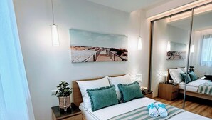 Une chambre moderne avec un plancher en bois, un lit avec des oreillers verts, une table de chevet avec une plante et un tableau représentant une scène de plage.