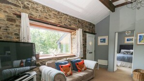 A cozy living room with a stone wall, a large window, a sofa with cushions, a TV on a stand, and a bed in the adjacent room. - The Old Barn (Lampeter)