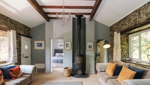 A cozy living room with a stone fireplace, a sofa with patterned cushions, and a large window with a view of greenery. - The Old Barn (Lampeter)