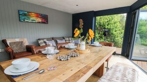 A dining area with a wooden table set for four, surrounded by leather chairs and a sofa. A painting is on the wall, and there's a window with a view of greenery. - The Old Barn (Lampeter)
