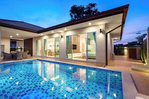 A modern house with a swimming pool, glass doors leading to a living area, and a well-lit exterior. - Luxury Pool Villa A18/3BR/8 Persons/BBQ (Pattaya City)