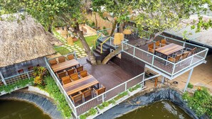 Een terras met meerdere niveaus, met houten tafels en stoelen, omgeven door weelderige beplanting en een rieten dak.